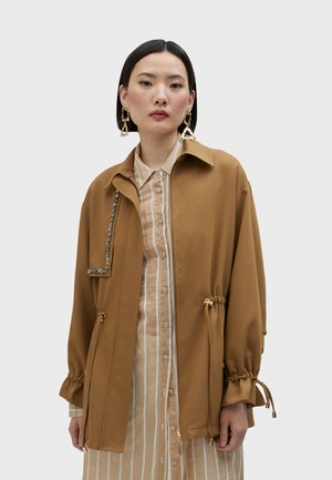 Giacca leggera marrone con chiusura a zip, dettagli con lacci in vita e un'applicazione decorativa in pietra su un lato.