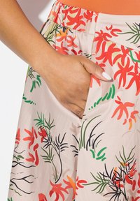 Jupe fleurie en tissu léger avec un fond beige et des motifs vifs rouges, verts et orange. Comprend une poche latérale.