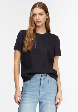 Woman with long light brown hair wearing a black short-sleeve shirt tucked into high-waisted light blue jeans against a plain background.