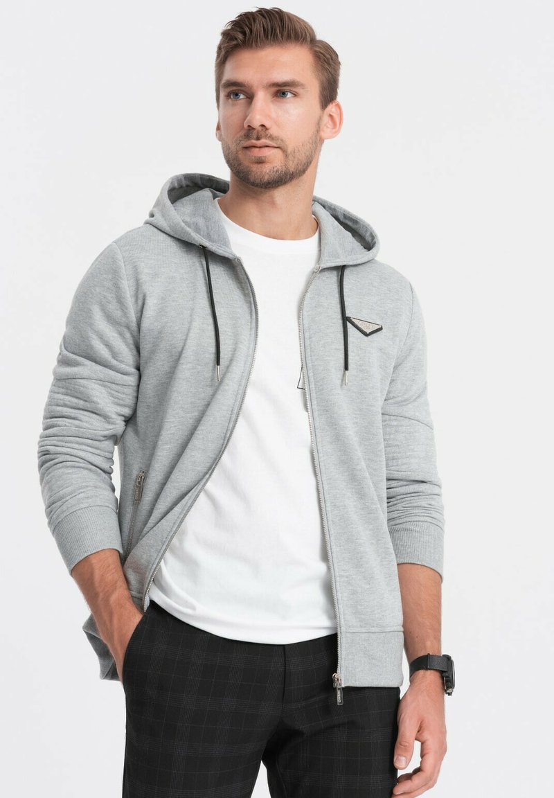 Felpa grigia con zip, cappuccio regolabile, tasche a marsupio e una piccola patch con logo. Indossata sopra una t-shirt bianca e abbinata a pantaloni a scacchi.