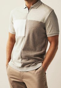 Polo gris clair avec un col zippé présentant un panneau gris foncé et une patch texturé blanc sur la poitrine. Tissu doux et décontracté.