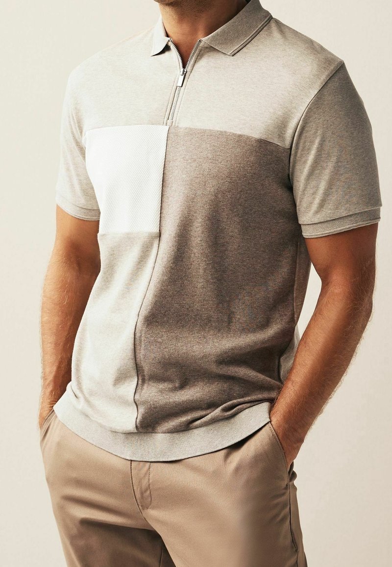 Polo gris clair avec un col zippé présentant un panneau gris foncé et une patch texturé blanc sur la poitrine. Tissu doux et décontracté.