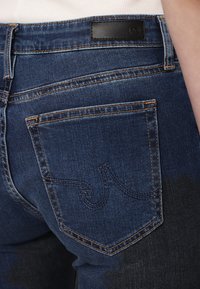 Jeans din denim de culoare albastră închisă au o buzunar din spate cu cusături sofisticate și un eticheta din piele neagră la talie, evidențiind o textură netedă.