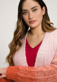 Neulejakku, jossa on gradienttisuunnittelu vaaleanpunainen runko ja kirkkaan oranssit hihat, esittelee tekstuurista, pörröistä kangasta.