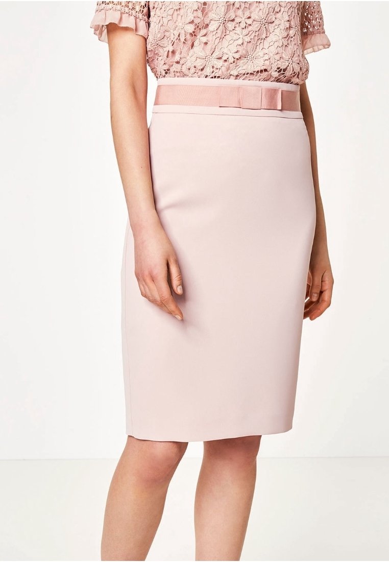 Femme portant une jupe crayon rose pâle longueur genoux avec une ceinture assortie et un chemisier en dentelle rose clair à manches courtes.