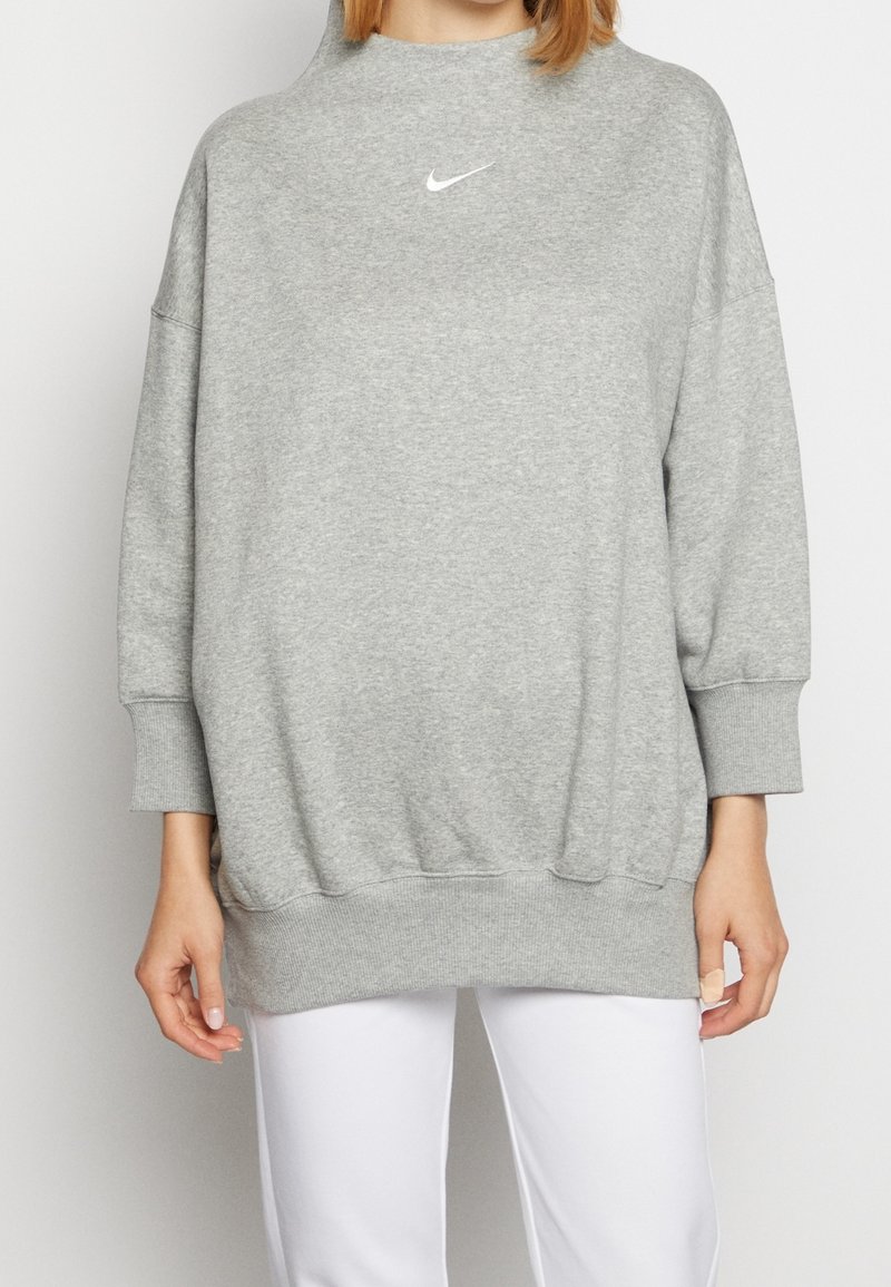 Sweatshirt Nike gris avec col montant, coupe oversize, poignets et ourlet côtelés. Présente un petit logo blanc sur la poitrine. Matière douce en mélange de coton.