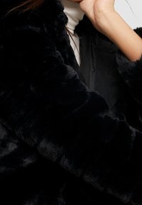 Cappotto in pelliccia sintetica nera con una texture morbida, caratterizzato da un fit leggermente oversize e un rivestimento interno liscio, visibile nell'area aperta.