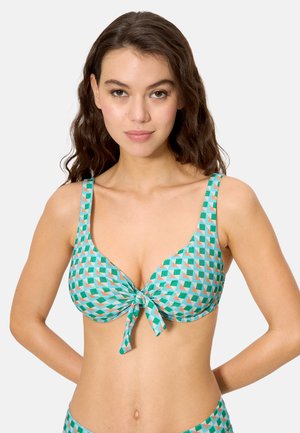 Joven con cabello castaño ondulado que lleva un top de bikini con patrón geométrico en verde y naranja, con un lazo frontal, contra un fondo claro y liso.