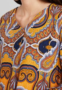 Nærbillede af en farverig bluse med paisleymønster i gul, orange, blå og hvid med V-udskæring, båret af en person med langt hår.