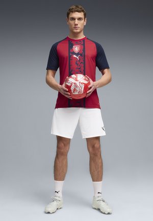 Mann in rotem und marineblauem Fußballtrikot und weißer Hose hält einen rot-weißen Fußball und steht vor einem grauen Hintergrund.