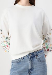 Sudadera blanca con bordado floral en las mangas, con cuello redondo, corte relajado y textura suave. Combina con jeans de color azul claro.
