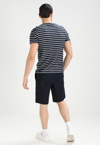 Randig marinblå och vit kortärmad t-shirt kombinerad med marinblå shorts. Vita strumpor synliga, bärs med vita sneakers. Neutral bakgrund.