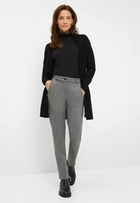 Cardigan lungo nero e maglione dolcevita abbinati a pantaloni grigi su misura e stivaletti neri; texture lisce e design semplice.