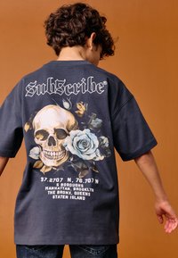 Sötét tengerészkék pamut póló, amelyen egy nagy koponya látható rózsák között, hátul pedig "Sub5cribe" felirat és koordináták vannak nyomtatva.