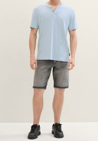 Un hombre se encuentra de pie con una camisa de manga corta azul claro, pantalones cortos grises y zapatillas negras, frente a un fondo neutro.