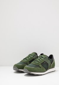 Scarpe sportive verdi in suede e mesh, con accenti neri, lacci rotondi e suola in gomma grigia. Logo evidente sul lato.