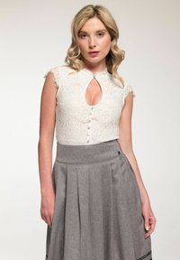 Vrouw met blond golvend haar, gekleed in een witte kanten mouwloze top met knopen en een sleutelgat uitsnede, gecombineerd met een hoog-geplaatste grijze geplooide rok.