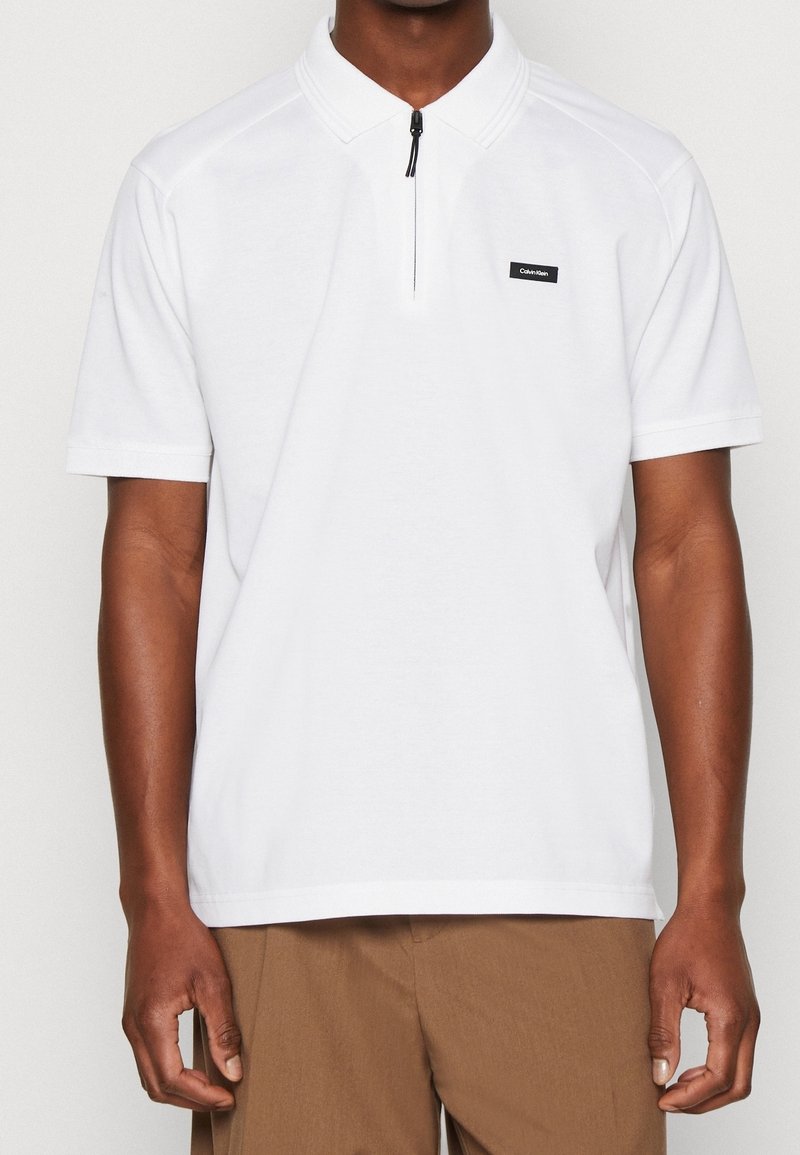 Polo blanc avec un col zippé, des manches courtes et une petite étiquette logo noire. En coton, avec une texture lisse.
