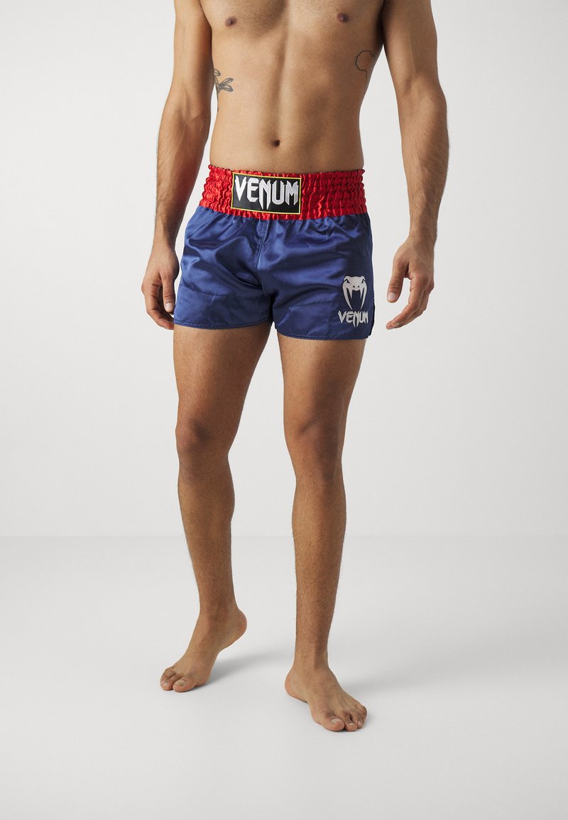 Pantalones cortos de satén azul con una banda elástica roja que presenta un logo. El diseño incluye un gráfico blanco en el muslo izquierdo.