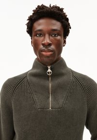 Junger Mann mit kurzem Afro-Haar, der einen olivgrünen gerippten Pullover mit einem halben Reißverschluss-Kragen trägt und direkt nach vorne schaut.