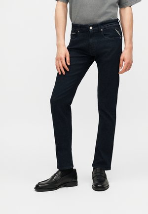 Άνδρας που φοράει σκούρα στενά τζιν, γκρι μπλουζάκι, μαύρες κάλτσες και μαύρα δερμάτινα μοκασίνια, στέκεται μπροστά σε ένα απλό ανοιχτό φόντο.