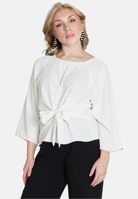 Blusa bianca con scollatura rotonda, maniche a tre quarti e dettaglio con nodo frontale. Texture liscia, vestibilità leggermente rilassata, abbinata a pantaloni neri.