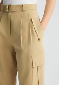Pantalons beiges avec des plis, dotés de grandes poches latérales, de détails en bouton et d'une ceinture assortie avec une boucle en métal rond.