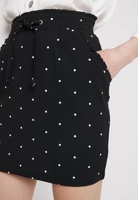 Falda negra con lunares blancos, cintura elástica con cordón, detalles con volantes a lo largo de la cintura y bolsillos laterales.