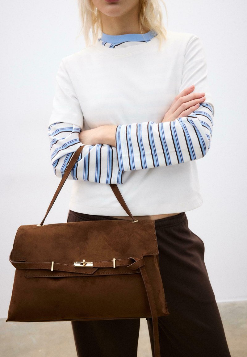 Personne en haut blanc avec manches longues à rayures bleues tient un grand sac à main en daim marron avec fermoir doré, debout contre un fond uni.