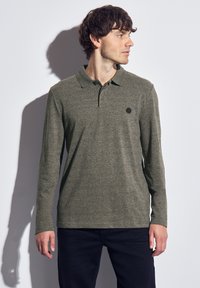 Polo à manches longues en vert olive, fabriqué en tissu texturé. Il présente un col classique et un petit logo noir sur la poitrine.