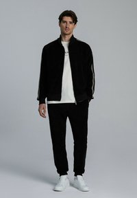 Czarny welurowy dres z białymi paskami po bokach, kurtka na zamek, spodnie z mankietami oraz białe trampki. Model stoi na neutralnym tle.