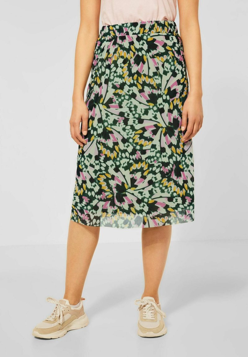 Woman wearing a knee-length skirt with green, pink, yellow, and black abstract floral pattern, paired with beige sneakers and a light top.