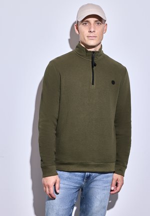 Oliven grøn pullover med kvart lynlås, struktureret stof og sort logo i accent. Sammen med lyseblå jeans. Justerbar krave.
