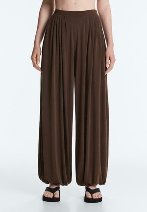 Braune locker plissierte Hose mit gerafften Säumen, getragen mit schwarzen Plateau-Sandalen bei einem barfüßigen Model, das steht.