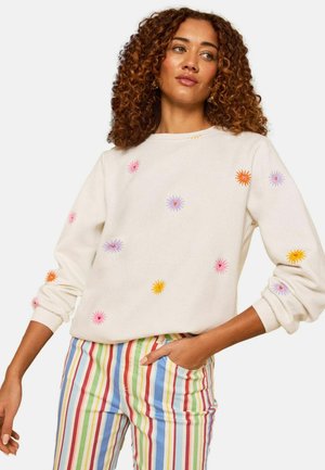 Kiharate jaustega naine, kes kannab valget pusa värviliste tikitud lilledega ja kõrge vöökohaga pükse, millel on vertikaalsed vikerkaare triibud.