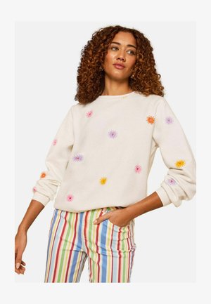 Femeie cu păr creț purtând un pulover alb cu flori brodate colorate și pantaloni cu talie înaltă și dungi verticale în culorile curcubeului.