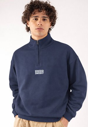 Mladý muž s kučeravými vlasmi oblečený v tmavomodrej fleecovej mikine so zipsom do polovice s logom "Koutko Society", béžové nohavice, stojaci so zdvihnutými rukami v vreckách na bielom pozadí.