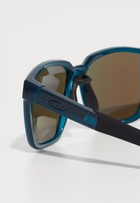 Gafas de sol con montura transparente azul y lentes marrones. Las características incluyen almohadillas nasales de goma y una varilla negra con un detalle de logo.