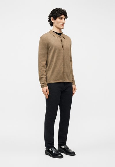 Άνδρας με σγουρά μαλλιά που φοράει καφέ μακρυμάνικο πουκάμισο με κουμπιά, μαύρο παντελόνι και μαύρα δερμάτινα μοκασίνια, στέκεται μπροστά σε μια απλή φόντο.