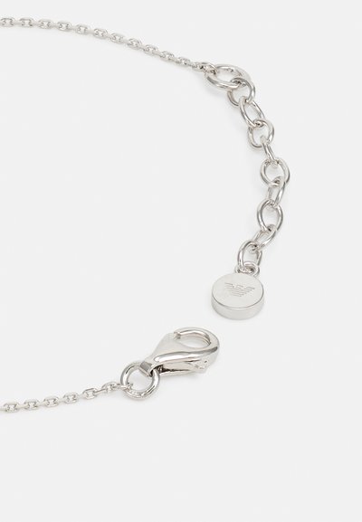 Silberne Kettenarmband mit ineinandergreifenden Gliedern, einem Karabinerverschluss und einem runden Anhänger mit Logo. Glatte Metalloberfläche, modernes Design.