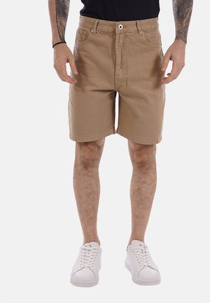 Uomo con pantaloncini beige fino al ginocchio, sneakers bianche e camicia nera, in piedi con le braccia rilassate ai lati.