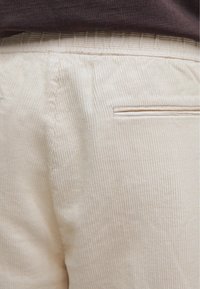 Close-up of beige corduroy pants with elastic waistband and a horizontal back welt pocket, worn with a dark brown top.
