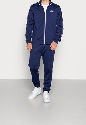 Homme portant un survêtement Nike bleu marine avec des bandes blanches, une fermeture éclair blanche et des baskets blanches, debout devant un fond uni.