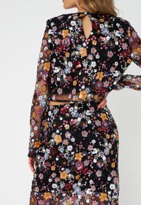 Abito nero floreale con maniche lunghe in tessuto trasparente, caratterizzato da vivaci fiori ricamati in viola, arancione e bianco, e un dettaglio a chiave sul retro.