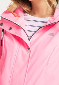 Rosa vattentät jacka med dragkedja och tryckknappsstängning, med en gråfodrad huva och en paraplylogotyp på bröstet. Randig skjorta under.