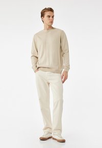 Suéter de punto beige con puños y dobladillo acanalados, combinado con pantalones rectos de un beige claro. El calzado presenta acentos en blanco y marrón.