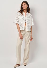 Witte t-shirt met korte mouwen en oogjespatroon, gecombineerd met lichtbeige textuurbroek en platte schoenen. De broek heeft een rechte pasvorm en knoopsluiting.