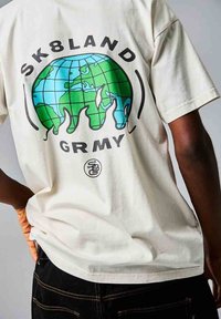 Camiseta de algodón color crema con un gráfico de un globo y llamas. El texto dice "SK8LAND" arriba y "GRMY" debajo del diseño.