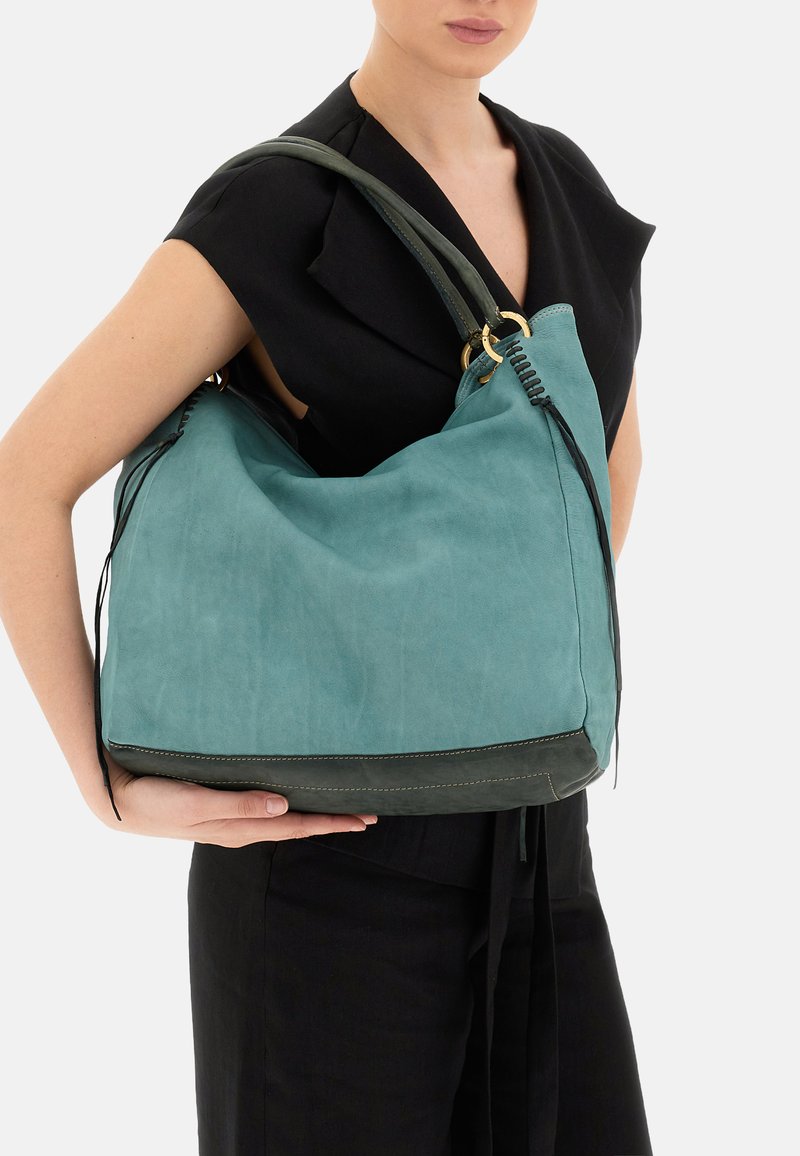 Borsa hobo in camoscio turchese con base in pelle verde scuro, dotata di accessori dorati e dettagli di cucitura nera, tenuta da una persona in una maglietta nera.