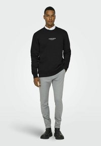 Schwarzes Baumwoll-Sweatshirt mit weißem Drucktext, kombiniert mit hellgrauen, schmal geschnittenen Jogginghosen und schwarzen Stiefeletten. Schlichter Hintergrund.